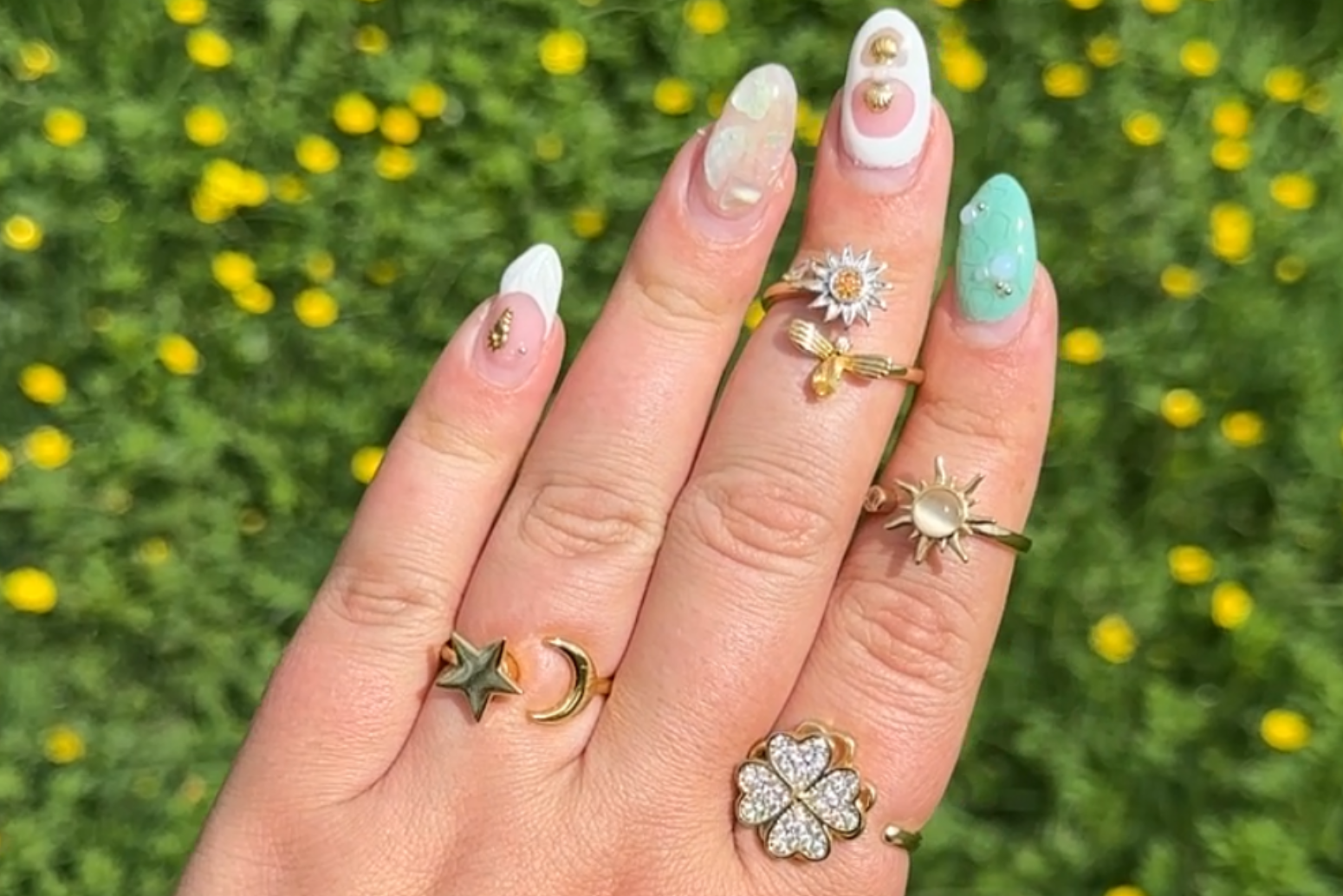 Hand with multiple rings and green nail polish against a grassy background